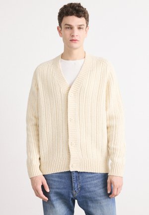 Cardigan en tricot côtelé crème avec un col en V, des boutons sur le devant et une coupe légèrement oversize, associé à un jean en denim bleu.