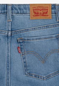 Pantalones vaqueros de mezclilla azul claro con una etiqueta de cuero con gráficos rojos, costuras amarillas y una pequeña pestaña roja en el bolsillo trasero.