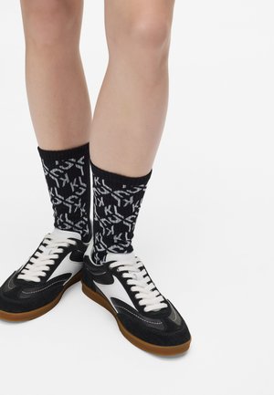 Mustvalged madalate tennisekatega kingad, mille pealispind on nahast ja nabuknahast ning mis on varustatud kummist tallaga. Kombineeritud mustade sokkidega, millel on graafiline muster.