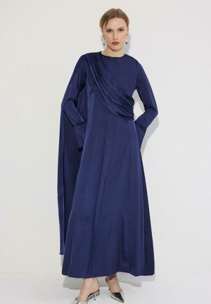 Vrouw die een lange marineblauwe jurk draagt met gedrapeerde stof over één schouder, zilveren puntige hakken en kleurrijke hangende oorbellen, staande tegen een witte muur.