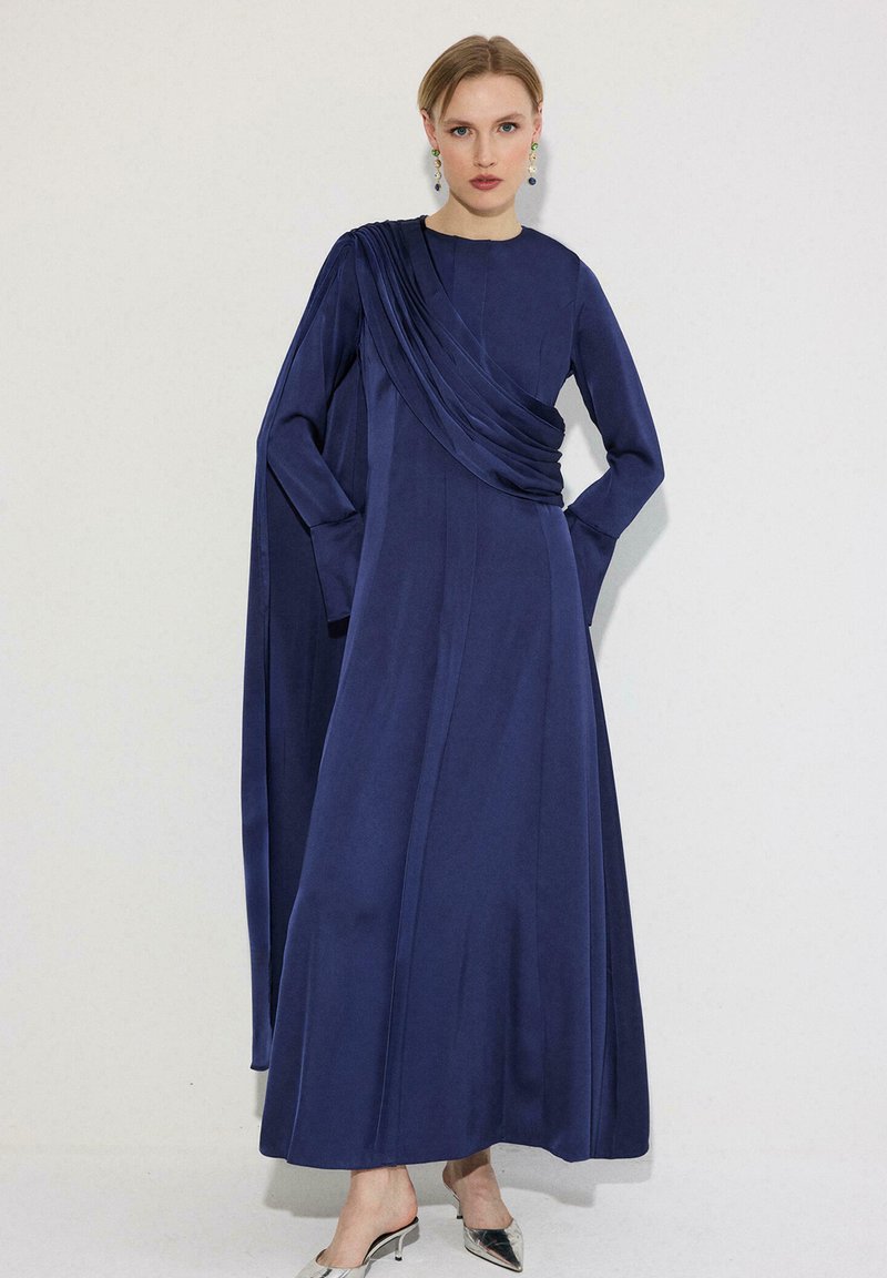 Γυναίκα που φοράει ένα μακρύ φόρεμα σε σκούρο μπλε με υφάσματα που πέφτουν πάνω σε έναν ώμο, ασημί μυτερά ψηλοτάκουνα και πολύχρωμα κρεμαστά σκουλαρίκια, στέκεται μπροστά σε λευκό τοίχο.