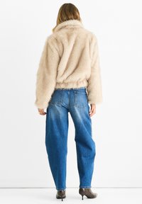 Woman wearing beige faux fur jacket, high-waisted blue jeans, and patterned heeled ankle boots, standing facing away on white background.