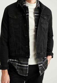 Zwarte denim jas met twee borstzakken en knoopsluiting, gelaagd over een witte geribde shirt en een zwart-grijze geblokte flanellen shirt.