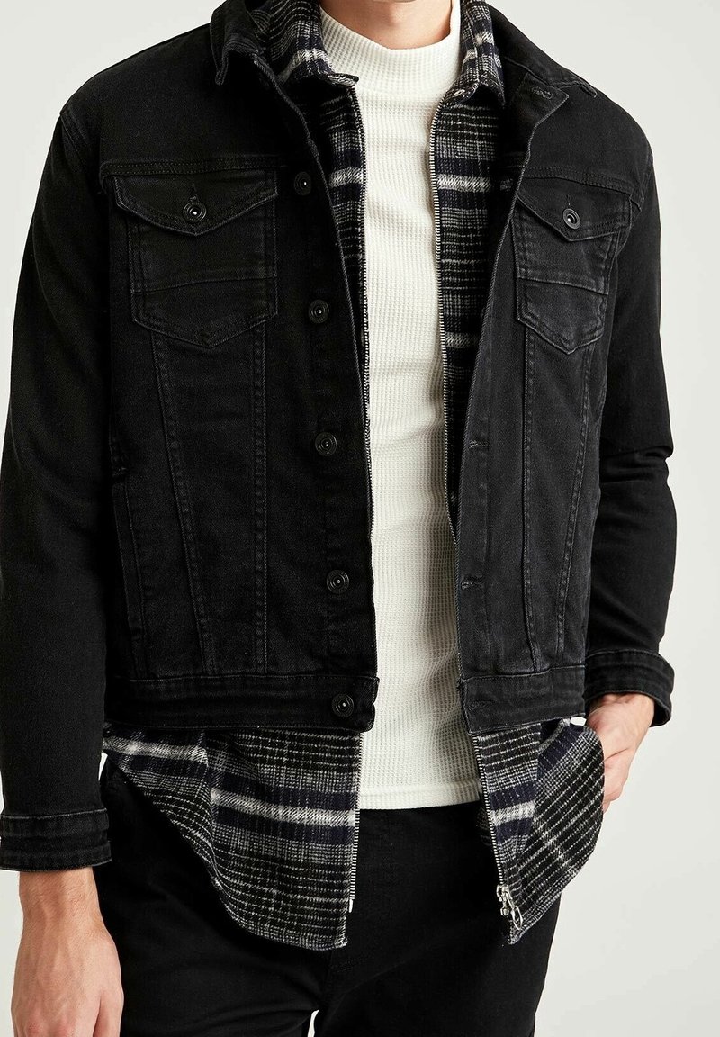 Schwarze Jeansjacke mit zwei Brusttaschen und Knopfverschluss, darüber getragen ein weißes Rippenshirt und ein karierter Flanellhemd in Schwarz und Grau.