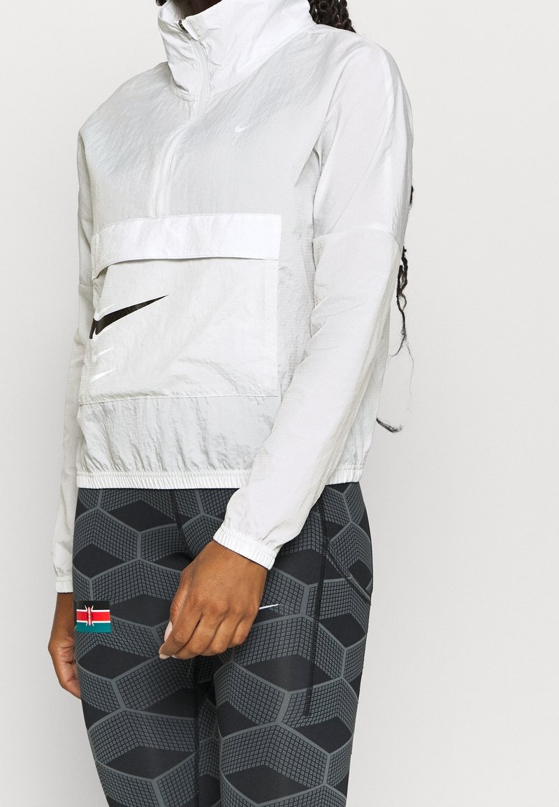 Veste légère Nike à demi-fermeture éclair blanche avec une poche avant. Associée à des leggings noirs à motif avec un écusson du drapeau kényan.