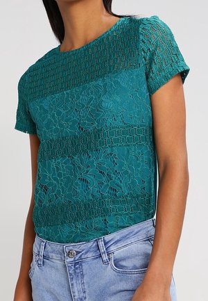 Sininen pitsinen lyhythihainen paita, jossa on kukka- ja geometrisia kuvioita, yhdistetty vaaleansinisiin farkkuihin.