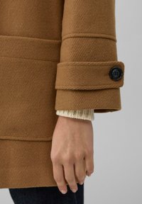 Hand, die einen beigen gestrickten Ärmel und einen braunen Mantelärmel mit schwarzen Knopfdetails trägt, liegt neben einer dunkelblauen Hose.