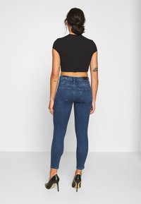 Dark blue skinny jeans in denim with back pockets, paired with a black cropped top and black high-heeled shoes. Visible tattoos on arms.