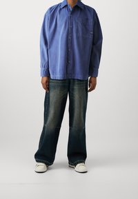 Blå oversized skjorta i bomull med framficka, matchad med mörkblå vida jeans och vita sneakers, med blekta texturer och en avslappnad design.