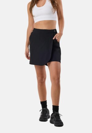 Kvinde iført en hvid sports-bh, sort atletisk skort med knappedetalje, sorte sokker og sorte løbesko, stående med den ene hånd i lommen.