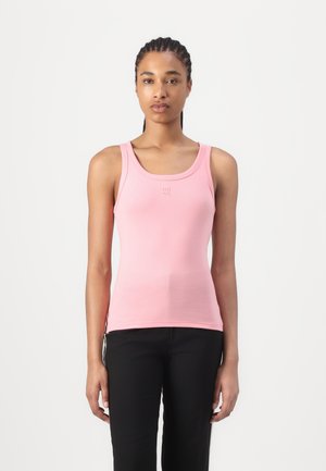 Lyserød ærmeløs tanktop med rund halsudskæring. Har et diskret logo på brystet. Materialet er glat og tætsiddende, kombineret med sorte bukser.