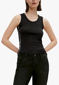 Top sin mangas de canalé negro con cuello redondo y detalles laterales con texto blanco. Combina con unos pantalones vaqueros negros de cintura alta.