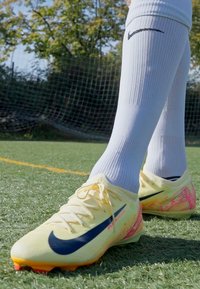 Žlté futbalové kopačky Nike s modrým logom, s textúrovaným povrchom a ružovými detailami. V páre s bielymi ponožkami po kolená.