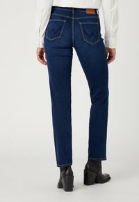 Donkerblauwe denim jeans met een rechte snit, zichtbare achterzakken met stiksels en een leren label op de tailleband. Zwarte haklaarzen.