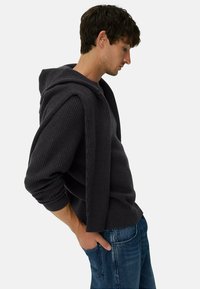 Donkergrijze geribbelde hoodie met een relaxed fit. Kenmerkt zich door een capuchon en gelaagd ontwerp, gedragen over een shirt, in combinatie met blauwe spijkerbroek.