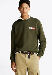 Sudadera de color verde oliva con puños y cuello acanalados, que presenta un parche de logo en blanco y rojo. Combinada con pantalones beige y un cinturón marrón.