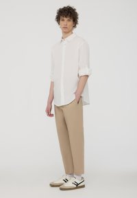 Camicia bianca a maniche lunghe con bottoni, abbinata a pantaloni beige a sigaretta e sneakers grigie con strisce nere, su uno sfondo semplice.