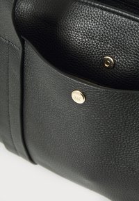 Sac en cuir noir à texture granuleuse avec une poche avant, doté d'une fermeture par bouton pression doré et d'un détail de logo embossé. La surface texturée est visible.