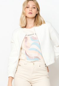 Bela teksturirana bomber jakna z zadrgo, nosena čez svetlo bež majico z grafiko, ki prikazuje pastelne valove. Ujemajoče lahke hlače.