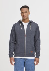 Sweat à capuche zippé bleu marine en tissu doux, avec un cordon de serrage à la capuche, des poches avant et un patch en cuir, associé à un jean bleu clair.