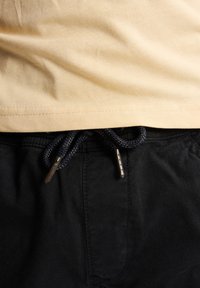 Beige katoenen t-shirt dat in zwarte broeken is gestoken met een gevlochten marineblauwe koord. Zilveren hardware op het koord en nette stiksels zijn zichtbaar.