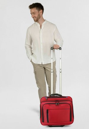 Junger Mann im weißen Hemd und beigen Hosen hält den Griff eines roten Rollkoffers, schaut nach unten und lächelt.