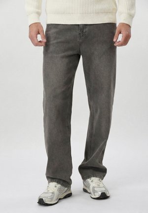 Mężczyzna ubrany w wyblakłe czarne jeansy o prostych nogawkach, biały dziergany sweter i białe trampki na jasnym, jednolitym tle.