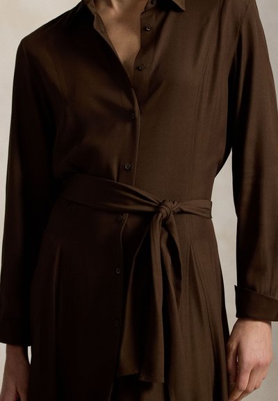 Καφέ μακρυμάνικο φόρεμα με κουμπιά και δέσιμο στη μέση, φορεμένο από άτομο με ένα χέρι ορατό στο πλάι.