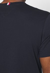 Camiseta azul marino con cuello redondo, mangas cortas y una pequeña franja roja, blanca y azul en la parte posterior del cuello. Textura suave.