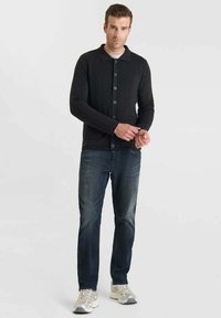 Uomo in cardigan nero a bottoni, jeans blu scoloriti e sneakers grigie che si trova davanti a uno sfondo chiaro e semplice.