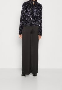 Top lungo a maniche lunghe nero con paillettes, colletto alto e schiena con keyhole; abbinato a pantaloni larghi in satin nero e scarpe con tacco alto.