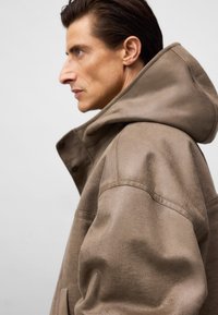 Brązowa kurtka z kapturem o luźnym kroju, z teksturowanej tkaniny, widocznymi szwami i zapięciem na zatrzaski z przodu. Ujęcie z profilu.