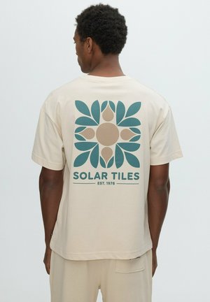 Hombre que lleva una camiseta beige con diseño floral verde y marrón claro y texto "Solar Tiles Est. 1976" en la espalda.