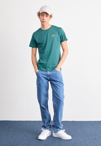 Zelena majica s kratkim rukavima s malim logotipom, uparena s svijetloplavim trapericama i bijelim tenisicama. Model nosi svijetlu kapu.