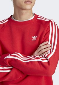 Felpa rossa con logo Adidas bianco sul petto e tre strisce bianche lungo ogni manica, realizzata in tessuto morbido con vestibilità comoda.