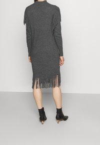 Femme portant une robe en tricot gris foncé jusqu'aux genoux, ornée de franges sur les manches et l'ourlet, assortie à des bottes noires à talons courts.