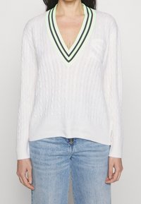 Weißer, kabelgestrickter Pullover mit einem tiefen V-Ausschnitt, der grüne und schwarze Streifen am Saum und ein Logo auf der Brust aufweist. Lange Ärmel und entspannte Passform.