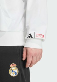 White sweatshirt with black Adidas stripes and a red Marvel logo, paired with black pants featuring the Real Madrid emblem in color.