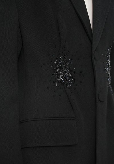 Černé sako s texturovaným povrchem, zdobené složitým korálkováním a ozdobami na ramenou. Zapínání na jeden knoflík a detail kapsy.