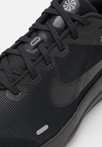 Črne športne čevlje z teksturirano mrežasto zgornjim delom, ki imajo sivo vezalke, gladek Nike logotip in subtilne odsevne detajle.