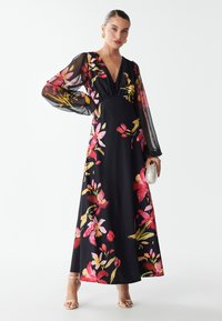 Abito maxi floreale nero con maniche lunghe trasparenti, scollatura a V profonda, caratterizzato da vivaci motivi floreali rosa, giallo e verde. Tiene una piccola pochette argentata.