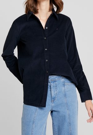 Personne portant une chemise noire à manches longues boutonnée et un jean taille haute bleu clair sur un fond uni.