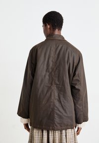 Veste marron, oversize, à manches longues avec une texture lisse et une couture au dos. Équipée d'un col montant et de manches légèrement retournées.
