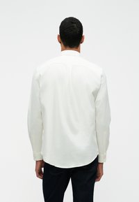 Camisa de manga longa, branca, feita de algodão, com um hem curvado, punhos com botões e um design de yoke nas costas sem padrões visíveis.