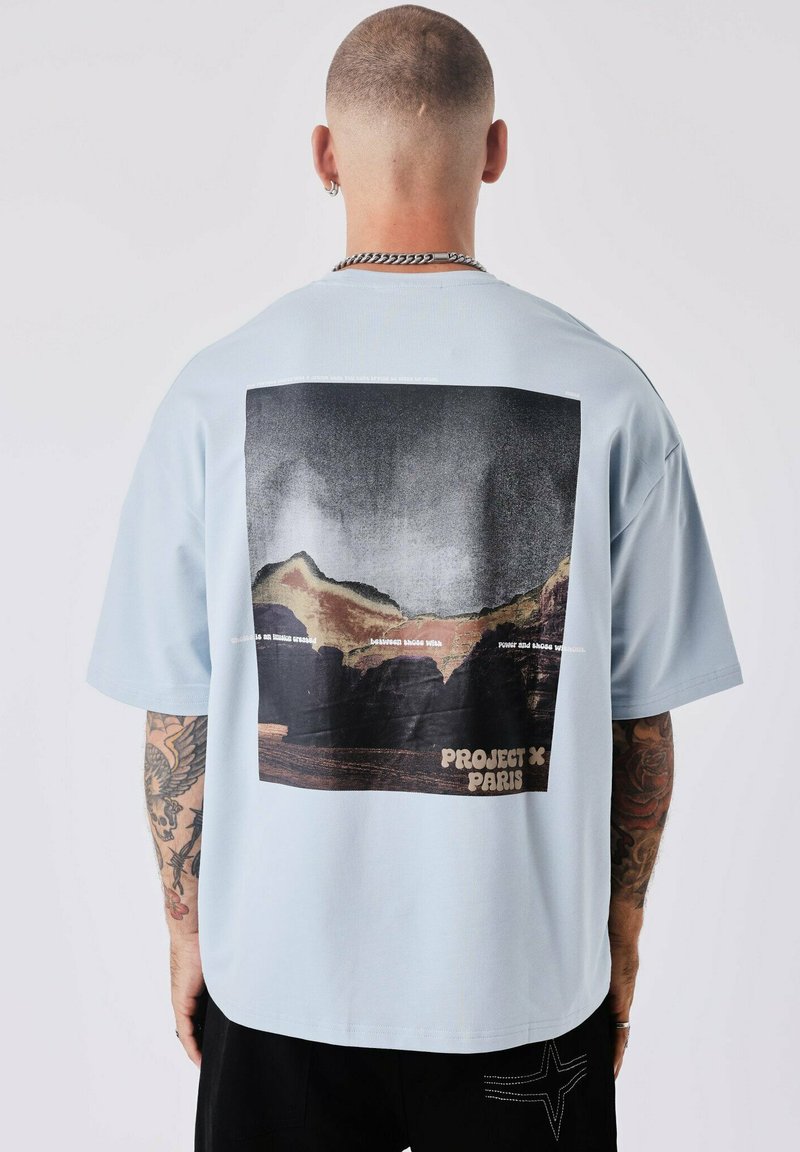T-shirt surdimensionné bleu clair avec un grand imprimé graphique noir de montagnes et du texte au dos. Manches courtes, en coton.