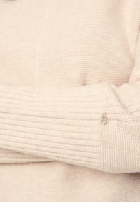 Hellbeiges Sweater mit gerippter Textur. Enthält einen metallischen Anhänger mit glitzernden Akzenten, der sich in der Nähe des Bündchens des Ärmels befindet.