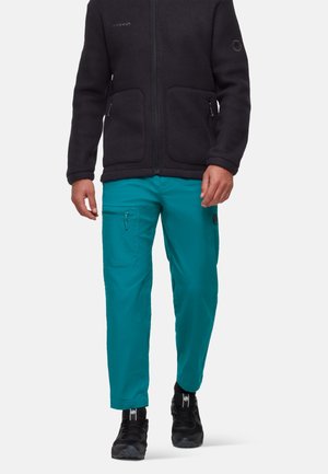 Uomo che indossa una giacca in pile nera e pantaloni cargo turchese con scarpe da ginnastica nere, in piedi davanti a uno sfondo uniforme.