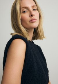 Čierny textúrovaný top so širokým dizajnom ramien, vyrobený z mäkkej látky a priliehavého strihu. Modelka má svetlo blond vlasy a neutralne nalíčenie.