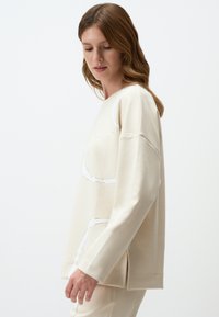 Creme sweatshirt met een ontspannen pasvorm, voorzien van witte diagonale naden en zijsplitten. Zacht stof met een gladde textuur, ronde halslijn.