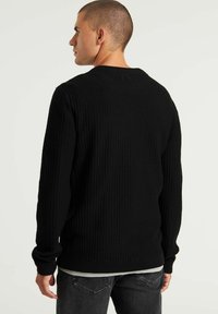 Maglione nero a maglia con maniche lunghe, tessuto strutturato, scollo a girocollo e polsini a costine; indossato sopra una camicia chiara con jeans grigi.